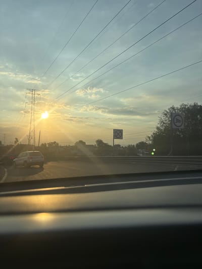 The image, taken from inside a vehicle, shows a view of what appears to be a highway in Tultitlán, Mexico. The sun is low on the horizon, causing a bright glare. Power lines and a transmission tower are prominent on the left side of the frame. Traffic signs are visible on the side of the road, including one that indicates lane merging, and another with a symbol indicating prohibited entry. Cars are present on the road. The scene suggests an early morning or late afternoon setting. The inside of the vehicle is visible on the right side of the image.