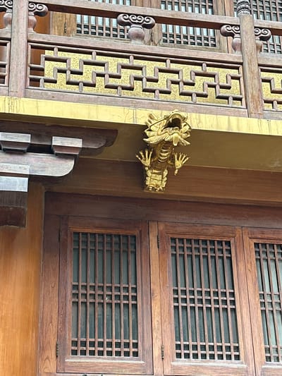 A close-up, eye-level shot features the facade of a building with traditional Chinese architectural elements in Jing'an District, China. The upper section of the building displays ornate wooden latticework with a golden decorative panel behind it, featuring a repeating geometric pattern. Below this, a prominent gold-colored dragon sculpture is affixed to the underside of a wooden eave, its head facing left with its mouth open. The lower portion of the image reveals two large wooden doors or windows, each with a grid-like pattern of vertical and horizontal wooden slats. The wooden elements are dark brown and appear aged. The wall to the left of the doors is a warm, orange-brown color. The lighting suggests it is daytime, with soft shadows indicating an overcast or diffused light source. There are no people or animals visible in the frame.