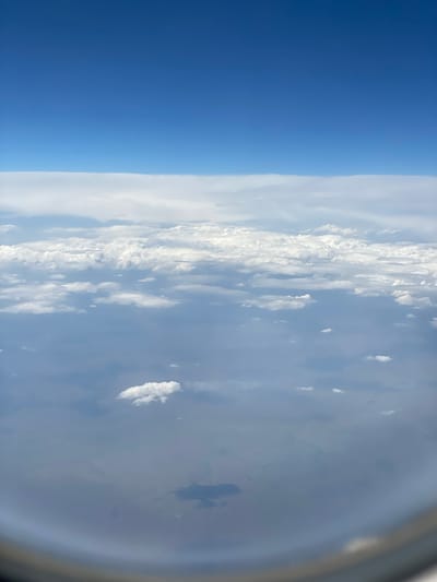 This is an aerial view, likely from an airplane, looking out a window at the sky and clouds. The top of the image is a clear, deep blue sky, transitioning to a layer of white, flat clouds. Below this layer, there are more scattered, puffy white clouds against a hazy, lighter blue sky. The lower portion of the image shows a view of the ground through a layer of haze or thinner clouds, with some landforms or bodies of water faintly visible. A curved, dark border at the bottom suggests the edge of an aircraft window. The scene is devoid of people or specific objects, focusing entirely on the atmospheric conditions and the distant landscape. The location in Медеу ауылдық округі, Kazakhstan, implies this view is over a rural or mountainous region, which might explain the faintly visible landforms.