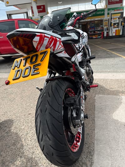 In Much Dewchurch, United Kingdom, a motorcycle with a custom paint job featuring red and white graphic designs is parked outdoors. The motorcycle's rear is prominently displayed, showcasing its yellow license plate which reads "MTO7 DOE". A black helmet with "X-804" branding rests on the motorcycle's seat. In the background, a red car is visible, along with a gas station featuring a "V-Power" sign and various fuel pumps. The ground is paved with asphalt and concrete.