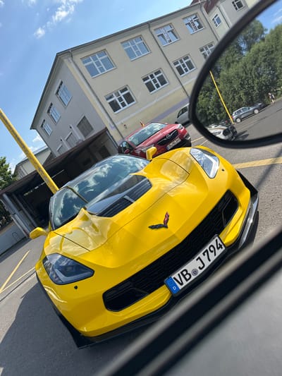 A bright yellow Chevrolet Corvette C7 is parked in a lot in front of a building in Alsfeld, Germany. The Corvette's license plate reads "VB J794". In the background, a red Opel Astra and other cars are visible. The building appears to be a school, with the letters "SPD" visible in one of the windows. The sky is blue with scattered clouds. The image is taken from a slightly elevated perspective, looking down at the Corvette. The lighting suggests it is daytime, likely afternoon, with the sun casting shadows.