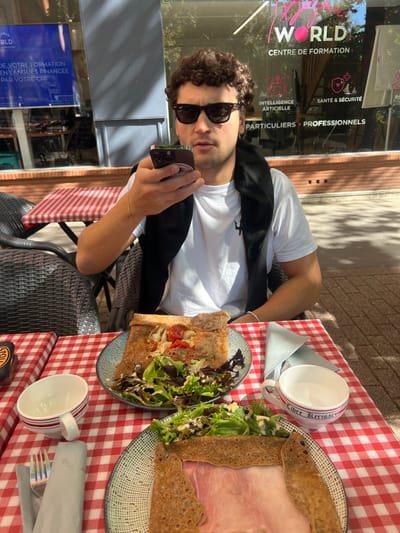 The image displays an outdoor cafe scene at 4 Rue des Anglaises in Orléans, France. A male individual with curly dark hair and sunglasses, dressed in a white t-shirt and a dark sweater draped over his shoulders, is seated at a table. He holds a dark-colored smartphone to his left ear or mouth, indicating an ongoing call or voice message recording. The table is covered with a red and white checkered tablecloth. On the table are two plates, each holding a savory crepe (galette) accompanied by a side salad with dressing. One crepe is topped with chicken and cherry tomatoes, while the other contains sliced ham. Two white ceramic cups with red stripes are present, one explicitly branded "L'ERE Kerisac." Silverware, including a fork and knife, is positioned to the left. The background features the glass facade of the "WORLD CENTRE DE FORMATION" building, with visible signage advertising "INTELLIGENCE ARTIFICIELLE," "SANTÉ & SÉCURITÉ," and "FORMATION EN LANGUES FINANCEE PAR VOTRE CPF." Wicker outdoor chairs are visible, and direct sunlight illuminates the scene.