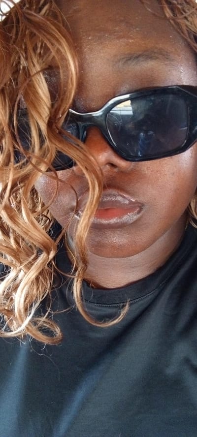 A close-up portrait shot captures a person with dark skin, likely in Alasia, Nigeria. The subject's face is partially framed by voluminous, light brown or blonde curly hair that cascades down the right side of their face, partially obscuring the forehead and cheek. They are wearing large, black, wrap-around sunglasses that completely cover their eyes. Reflections in the sunglasses suggest an indoor setting or the interior of a vehicle, possibly showing a steering wheel or dashboard.
The person's lips, which appear naturally full, have a slightly glossy finish. A plain black t-shirt is visible from the neckline downwards. The skin on the forehead and cheek areas shows natural texture. The image focuses on the person's face and upper chest, with the background out of focus. The lighting appears to be natural, consistent with daytime. There is no discernible activity other than posing for the photograph. No text is visible in the image.