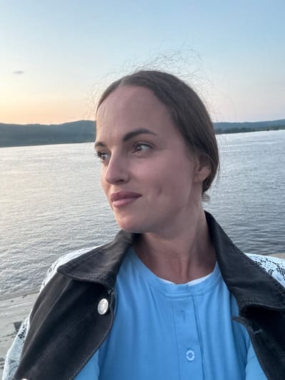 A medium shot captures a woman in the foreground, her face turned to the left as she looks out at a body of water under a pale sky. She has brown hair pulled back, fair skin, and dimples visible on her cheeks. She is wearing a light blue long-sleeved shirt with buttons down the front, and a dark jacket with white lace detailing on the sleeves is draped over her shoulders. The background consists of a wide expanse of water reflecting the muted light of the sky, and a distant, hazy shoreline with rolling hills. The scene is tranquil and appears to be set outdoors, possibly at dusk or dawn. The location is specified as Probuzhdenie, Russia.