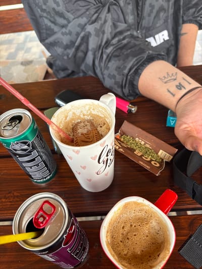The image shows a wooden table with various items placed on it. On the table, there are two mugs, one with a red handle filled with foamy coffee, and the other with pink hearts on it and also filled with a coffee-like beverage with a biscuit inside and a red straw in it. There are two open cans of energy drinks, one green with the brand name "HELL" visible and a red straw in a purple can. A pink lighter, a pack of OCB rolling papers with some loose material that resembles weed, a blue pack, and a black strap are also on the table. An arm with tattoos, including a crown and the numbers "1 of 1", rests on the table near the rolling papers. The person wearing a black patterned jacket is partially visible in the background. The scene is reminiscent of a table after people have been drinking coffee and energy drinks, possibly preparing to roll something. There is no indication that this is in Polena, Bulgaria.