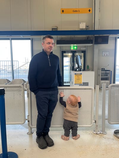 The image depicts an indoor scene within an airport gate area, specifically Gate 1, at what is likely Frankfurt-Hahn Airport in Lautzenhausen, Germany.
An adult male, approximately 30-40 years old, with short dark hair and light grey temples, stands behind a perforated light grey metal barrier. He is wearing a dark blue quarter-zip sweater, dark blue trousers, and dark grey low-cut boots. His left hand is in his pocket, and his gaze is directed towards the right of the frame.
To his right, an infant or toddler stands facing away from the camera, also behind the barrier. This child has reddish-blonde hair and wears a dark long-sleeved top, dark trousers, a light brown sleeveless vest, and patterned soft-soled shoes. Both of the child's hands are raised and placed on the top edge of the metal barrier.
In the background, large windows to the left reveal an outdoor airfield with metal structures. To the right, a white wall features a yellow overhead sign with black arrows and text reading "Gate 1". Below this, a glass door with a green emergency exit sign above it is visible. Adjacent to the door is a white self-service kiosk with a black monitor and scanner on top. A yellow and white poster on the kiosk partially reads "HOT DRINKS", "Attention Passengers", and "RYANAIR GROUP". Various wall-mounted fixtures, including electrical boxes and vents, are present. Further to the right, a small desk with a microphone and paper holder is seen, along with two "No Smoking" and "No Vaping" circular signs on the wall. The floor is a light, neutral color.