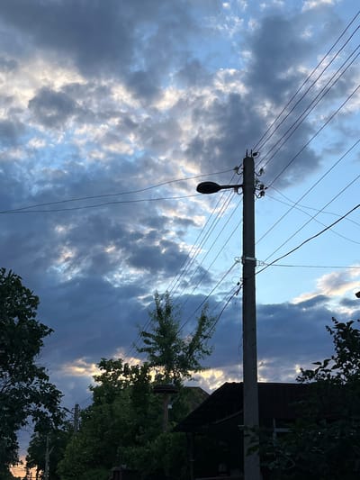 The image depicts an outdoor scene at dusk in Nikolayevka, Russia. A concrete utility pole with a streetlamp and multiple electrical wires dominates the right side of the frame. The sky is filled with dramatic, dark gray clouds interspersed with patches of lighter, possibly illuminated by the setting sun. Silhouettes of trees and the roof of a building are visible in the lower part of the image. The overall atmosphere is somber and transitional, indicating the end of daylight. There are no people or animals clearly visible in the scene.