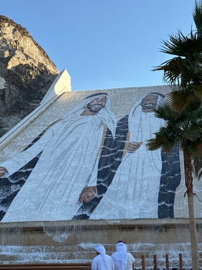 The image depicts the Hatta Dam in Hatta, United Arab Emirates. The sloped face of the dam features a large-scale mural rendered in a mosaic or textured style, portraying two smiling male figures. Both figures wear traditional white Emirati attire, including ghutras and kanduras. Water continuously cascades down the entire mural, creating a dynamic, textured visual effect as it flows into a tiered basin at the dam's base. The figure on the left extends his left arm downward, and the figure on the right extends his right arm downward. To the left of the dam, a rugged, sunlit rocky mountain range rises against a clear blue sky, which dominates the upper frame. A green palm frond is partially visible on the far right, obscuring a small portion of the dam. In the lower foreground, two individuals are observed from behind, positioned at the dam's base. Both wear traditional white Emirati clothing, including ghutras, and face the dam, appearing to observe the mural and water flow. Partial wooden structures, possibly a bench and bollards, are visible in front of and to the right of the individuals.