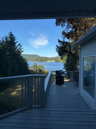 The image displays a clear daytime view from a grey wooden residential deck overlooking Shawnigan Lake in Area B, Canada. The deck features a matching railing, incorporating clear glass panels on its right side. A black propane barbecue grill stands on the deck, and two dark patio chairs are visible to its right, positioned near a light-colored building wall with a large, reflective window. A light blue/grey soffit panel is visible overhead, forming the top frame. The midground shows the blue water of Shawnigan Lake, bordered by dense green coniferous trees along the shore and distant tree-covered hills. Several structures are discernible on the far side of the lake amidst the vegetation. The sky is bright blue with a single, elongated white cloud. No individuals are present, and no active interactions are depicted within the scene.