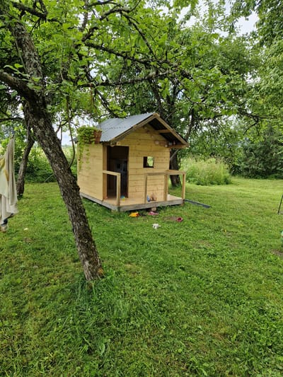 An outdoor, eye-level, medium shot captures a wooden playhouse situated on a grassy lawn beneath the canopy of a mature tree. The playhouse, made of light-colored wood, features a corrugated metal roof, a small window, and an open doorway leading to a porch with a railing. Toys, including a yellow duck, a white stuffed animal, and a pair of pink shoes, are scattered on the grass in front of the playhouse. A beige towel hangs from the trunk of the tree on the left side of the frame. The environment appears to be a rural or suburban backyard, likely in the city of Beja, Latvia, given the natural setting with abundant greenery and trees. The lighting suggests it is daytime.