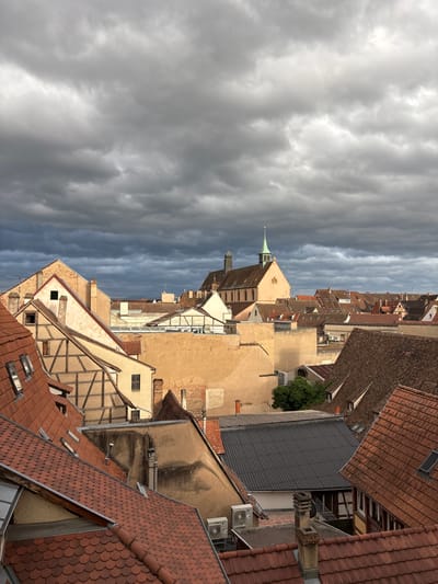 The image shows a view of rooftops in the historic district of Colmar, France, under a dramatic, overcast sky. The architecture features a mix of traditional Alsatian half-timbered buildings with exposed wooden beams and lighter-colored facades, alongside buildings with terracotta tiled roofs and plastered walls. A prominent church with a green steeple and a bell tower is visible in the background, its ochre-colored walls catching the light. The foreground is dominated by the red-tiled roofs of various buildings, some with visible dormer windows. The overall impression is one of dense urban development with a strong sense of history. There are no people or animals visible in the image. The environment appears to be a residential or mixed-use urban area.