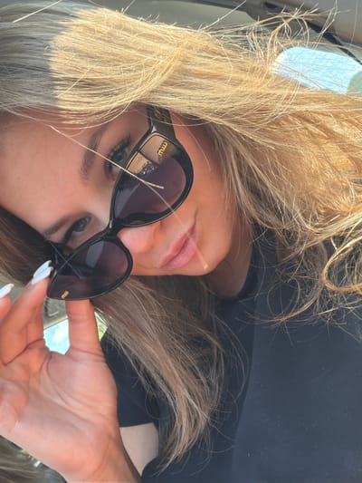 A close-up selfie captures a young woman with blonde hair and blue eyes, wearing black sunglasses. She is holding the sunglasses with her left hand, showing off her white-painted fingernails. Her lips are slightly parted as she looks towards the camera. The background is blurred, suggesting she is inside a vehicle, as indicated by the car's interior visible at the top of the image. The lighting suggests it is daytime, with sunlight filtering through her hair and casting shadows. The scene is set in Palje Brdo, Croatia.