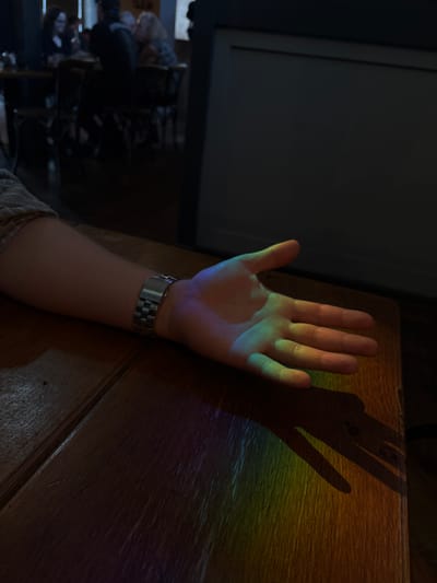 The image depicts a close-up view of a person's outstretched left hand, illuminated by a rainbow-colored light refraction, casting a shadow on a wooden table. The hand, appearing to be a Caucasian adult's, is positioned palm-upwards, with the fingers slightly spread. A silver wristwatch is visible on the wrist. The light source creating the rainbow effect is not directly visible in the frame, but it appears to be a directed beam of light passing through an object that causes refraction. The background is dark and out of focus, suggesting an indoor setting, possibly a dimly lit room. Faint figures of people can be seen in the far background, appearing to be seated at tables in what could be a restaurant or bar. There is no discernible indication of the location being in Ipswich, United Kingdom. The scene is static and captures a moment of light interaction with a human hand.