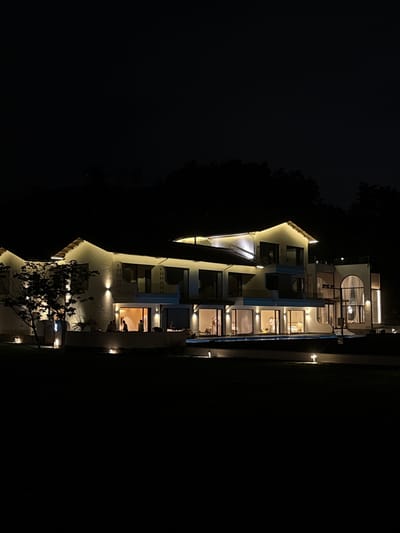 The image shows a multi-story building illuminated at night, situated in Jinhua, China. The structure appears modern with a combination of solid walls and large windows. Light emanates from within the building and from exterior lighting fixtures, suggesting residential or commercial occupancy. The surrounding environment is dark, with visible trees or vegetation to one side and a dark expanse on the other. No people are clearly visible in the image, though there are suggestions of figures inside the building through the lit windows.