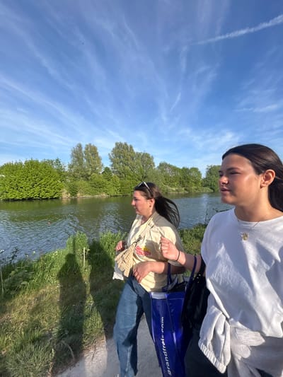 Two young women stand near the edge of a body of water, possibly a canal or river, in Marquette-lez-Lille, France. The upper woman wears a white shirt, and the lower woman wears a tan shirt and jeans. A blue bag with the text "grandfrais.com" is present. The environment includes green foliage along the water's edge and trees in the background. The sky is blue with scattered clouds.