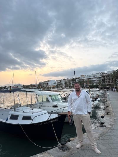 A man stands on a pier in Port d'Alcúdia, Spain, with a line of docked boats behind him and buildings and palm trees visible in the background. The sky is overcast with a hint of sunset colors peeking through. The man is wearing a white linen shirt and beige pants. He is holding a dark object in his left hand. The pier is paved with stone, and there are bollards for tying up boats. The boats are of various sizes, mostly white with some dark accents. The water appears calm.