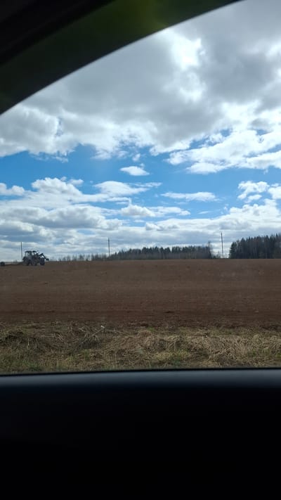 The image shows a landscape as seen from inside a vehicle, likely a car or truck. The sky is visible, with a mix of blue sky and white, puffy clouds. A field extends from the center of the frame to the right, transitioning from plowed earth to a strip of dry grass and vegetation. A line of trees can be seen in the distance. The edge of the vehicle's window frame appears on the left, and the vehicle's interior is visible on the right side. A tractor is located in the field.
The location is potentially Малая Уса, Russia, though this cannot be confirmed with visual data alone.