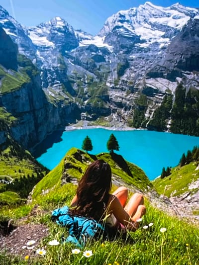 The image depicts a scenic mountain landscape with a person enjoying the view. The scene includes a rugged, snow-capped mountain range on the left, transitioning to a steep, green mountainside dotted with trees. A vibrant turquoise lake is visible in the middle ground, reflecting the bright sky. On the right side of the frame, a person with long, dark hair is lying in lush green grass, facing away from the camera. They are wearing shorts and a top, with a blue backpack beside them. Small white wildflowers are scattered throughout the grass. The perspective suggests the person is relaxing and taking in the breathtaking scenery. The lighting indicates it is daytime, likely sunny, given the bright colors and clear blue sky. While the image is stunning, it does not contain any visible text. The location context provided of Nijmegen, Netherlands, is inconsistent with the mountainous and alpine scenery shown in the image.