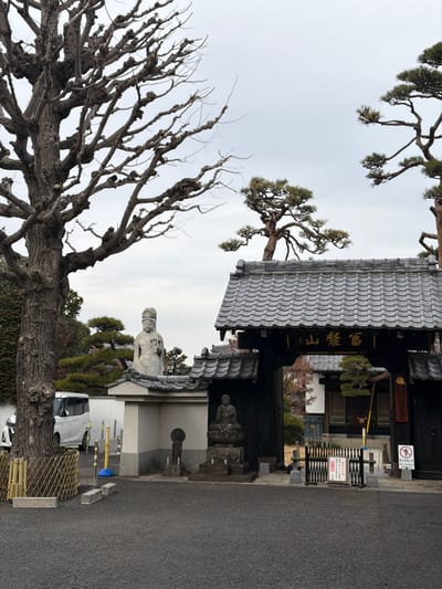 A traditional Japanese building with a tiled roof stands near a street. Ornate bare trees frame the left side of the image, contrasting with a pruned pine tree above the building. Several stone statues rest on the building's front, while signs hang near a gate. A gray sky contrasts with the building's dark facade.