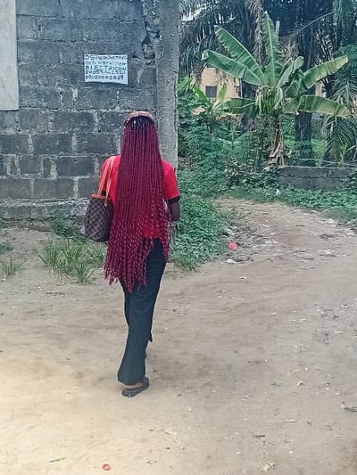 An individual is depicted walking away from the camera on a light brown dirt path in Ifite Awka, Nigeria. The person wears a red short-sleeved top, dark trousers, and dark slip-on sandals. Their back is to the viewer, revealing long, dark red braided hair extending to the waist. A brown patterned shoulder bag with a distinct monogram design is carried on the left shoulder. To the left, a weathered grey concrete block wall displays a white rectangular sign with black text advertising "DStv / Startimes GOTV Installer ELECTRICIAN" and contact numbers "07038662320" and "09020272869". The immediate surroundings feature sparse ground vegetation and debris on the path. To the right and in the background, dense green foliage, including prominent banana and palm trees, partially obscures a light-colored building with a visible window.