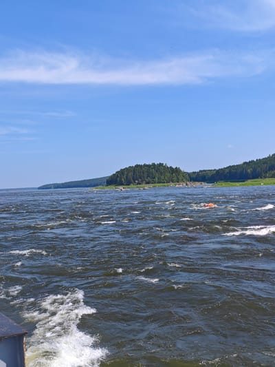 The image depicts a wide, fast-flowing river in the city of Porog, Russia. The water is dark and turbulent, with whitecaps and foam indicating a strong current. In the distance, a forested peninsula juts out into the river, with a line of trees along its edge. The sky is a clear blue, with a few wispy white clouds. On the river, a bright orange object, possibly a buoy or a piece of equipment, is visible amidst the waves. The foreground of the image shows a portion of a vessel, likely a boat or ship, from which the photograph was taken, with white water trailing behind it. The overall scene conveys a sense of natural power and the vastness of the riverine landscape.