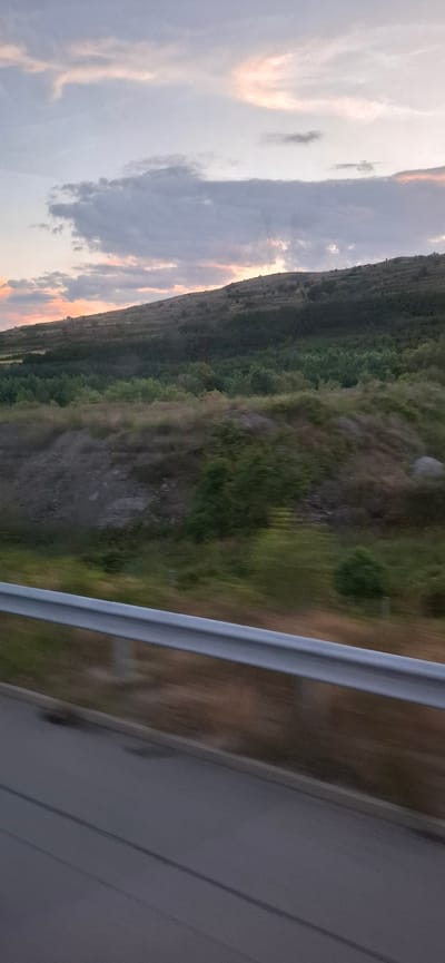 The image captures a blurred landscape scene, likely taken from a moving vehicle, with a focus on a rolling hill against a soft sky. The sky exhibits pastel colors, with hints of pink and orange from what appears to be a sunset or sunrise, and is dotted with scattered clouds in shades of gray and white. Below the sky, a sloping hill is visible, covered in vegetation that transitions from sparse, dry grass near the top to denser green foliage further down. The bottom of the image shows a portion of a road with a silver guardrail running horizontally across the frame. The blurriness suggests motion. The location is identified as Djerman, Bulgaria.