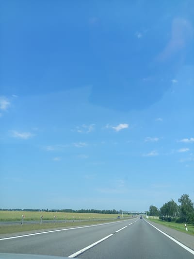 The image depicts a highway scene on a clear, sunny day in the vicinity of Валевачскі сельскі Савет, Belarus. The perspective is from a vehicle traveling on a multi-lane asphalt road. The road is bordered by white lines and guardrails with red and white markers. To the left of the road, a green field extends to the horizon, dotted with sparse trees and structures. To the right, a grassy area is followed by a line of trees. In the distance, a highway overpass can be seen, with a few vehicles on the road. The sky is a vibrant blue with scattered white clouds. There are no people visible in the image.