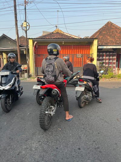 In Sugih Waras, Indonesia, four individuals are observed on motorcycles on a paved road. The foreground features a black Honda scooter with a license plate reading "3086 TFD." To the right of the scooter is a red motorcycle with a license plate "P 2905 HF." Behind the red motorcycle is another scooter with a license plate "W 6505 ZF." The fourth motorcycle is partially visible, behind the scooter with the "W 6505 ZF" license plate.
The individuals are wearing helmets and are either operating their motorcycles or waiting. One person on the black scooter wears a black jacket. The person on the red motorcycle has a backpack and is facing away from the camera. The person on the "W 6505 ZF" scooter is a woman wearing dark clothing with white stripes and patterns. The fourth individual is only partially visible.
The environment suggests a residential area with buildings featuring tiled roofs and orange gates. Utility poles and wires are visible in the background. The lighting indicates it is daytime, likely late afternoon due to the warm tones. The road surface appears worn.
