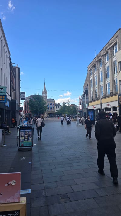 This image depicts a street scene in Leicester city centre, United Kingdom, likely on a sunny day, given the bright blue sky with scattered clouds. The perspective is from within a pedestrianized shopping area.
On the left side of the street, a white building with multiple windows dominates the foreground, with shopfronts visible below. One sign reads "KOKORO" and another partially obscured sign shows "GREGGS". Further down, a person is seated in a wheelchair. A black A-frame sign with an image of a gorilla and text that reads "SELL US YOUR PHONE!" is positioned on the pavement.
The centre of the image reveals a wide, paved pedestrian walkway with people walking in both directions. In the distance, a prominent clock tower with a spire stands tall, a focal point of the cityscape. Buildings of varying architectural styles line both sides of the street. On the right, a multi-story building with classical architectural elements and numerous windows is visible, with a prominent HSBC bank branch on the ground floor.
Several individuals are captured in motion: a man with a light-colored patterned shirt and a dark bag walks away from the camera; another person in a dark jacket and trousers walks towards the camera. Groups of people are seen dispersed throughout the area, some appearing to be shopping or socializing. The pavement is made of large, dark grey rectangular tiles.
The overall impression is of a lively urban environment, likely a main thoroughfare for shopping and leisure in Leicester.