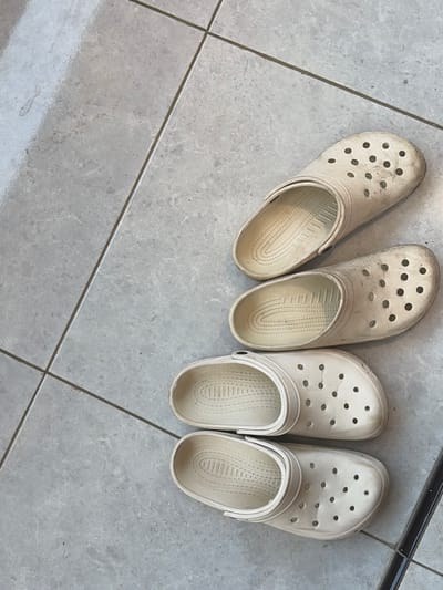 Four pairs of white clogs are arranged diagonally on a tiled floor. The clogs are worn and show signs of dirt and use. The floor is made of large, light grey tiles with dark grout lines. The lighting suggests an indoor environment. The scene is devoid of any people or any indications of activity.