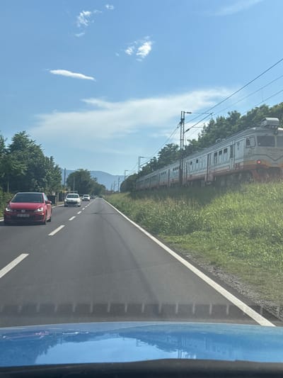 This image captures a daytime scene on a paved road, likely a highway, with a train track running parallel to it on the right. The road is marked with white lines and has a shoulder. Several cars are visible on the road, including a red car in the foreground on the left and white cars further ahead. A train is on the tracks, appearing to be in motion or stopped, with multiple carriages. The environment is rural or semi-rural, with lush green grass and trees lining the road and the train track. In the background, hills or mountains are visible, and the sky is blue with some scattered white clouds. The image appears to be taken from inside a vehicle, as indicated by the reflection on the blue surface in the foreground. The scene is consistent with a location in Bijelo Polje, Montenegro, a region known for its natural landscapes and transportation infrastructure.