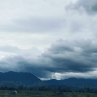 The image presents a wide, overcast landscape, likely captured during the day in Keureuweungkrueng, Indonesia. The scene is dominated by a dramatic sky filled with thick, textured clouds ranging in color from pale grey to deep, moody blue-grey, suggesting heavy cloud cover or an approaching storm. These clouds occupy the majority of the frame, creating a vast and somewhat somber atmosphere.
In the mid-ground, a range of dark mountains rises, their undulating peaks appearing as deep blue-grey silhouettes against the cloudy sky. Their forms are partially softened by atmospheric haze and the diffuse light, seamlessly blending with the darker clouds in places.
The foreground consists of a blurry, dark green expanse, likely a field or open land. A faint, light-colored shape is barely discernible in this area, possibly an animal, though its details are too indistinct to confirm. The overall color palette is composed primarily of cool tones, with blues, grays, and dark greens lending a serene yet powerful feel to the natural vista. There is no visible text in the media file.