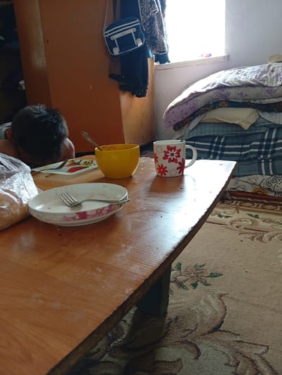 A low-angle, medium shot captures a close-up of a wooden table laden with dishes and a person sleeping. The table is positioned in a room with warm lighting, likely from a window on the right, which illuminates a stack of neatly folded blankets and bedding. To the left, a person, possibly a child, is seen sleeping with their face buried in their arms on the table. The table holds a yellow bowl with a spoon, a white floral-patterned plate with a fork, and a white mug adorned with red flowers, also containing a spoon. A plastic bag containing bread is also present. The background reveals a tall wooden cabinet and a patterned rug covers the floor. The scene appears to be in a home environment in Shelek, Kazakhstan.