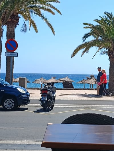 This image captures a vibrant coastal scene in Puerto Del Carmen, Spain, bathed in bright daylight under a clear blue sky. In the immediate foreground, part of a dark wooden table and a woven chair suggest a café or restaurant patio.
Beyond this, a paved road features a dark blue Fiat Panda partially visible on the left and a black scooter (license plate 2353 JGG) parked on the right. A yellow line marks the road. Adjacent to the road, a blue "No Parking" sign with a red diagonal slash is mounted on a pole. Below it, a white sign specifies "DE 8.00 A 15.00H Excepto Carga y Descarga Horario: Ma, Mi, Jue, Vi Máx. 30 Min Domingos y Festivos: Libre" (From 8:00 to 15:00, Except Loading/Unloading Hours: Mon, Wed, Thu, Fri Max. 30 Min, Sundays and Holidays: Free).
Between the road and the sea, a promenade or sidewalk stretches, shaded by tall palm trees on both sides of the frame. A man and a woman are walking from right to left on this promenade. The man wears a dark cap, dark top, and light shorts, while the woman is in a red top and dark pants with a white stripe. Their shadows are cast distinctly on the pavement, indicating strong sunlight.
In the background, the vast, calm blue ocean extends to the horizon. Closer to the shore, several straw umbrellas (palapas) are visible on what appears to be a rocky beach or coastal area, with a square concrete structure nearby. The overall atmosphere is relaxed and scenic, typical of a sunny Mediterranean or Canary Islands locale.