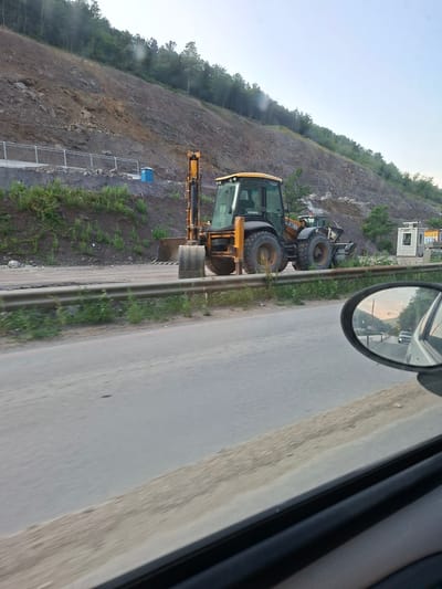 The image shows a backhoe loader parked on a construction site next to a road. The construction site appears to be on a hillside, with a steep, rocky slope behind it. There is a guardrail separating the road from the construction site. In the foreground, the reflection of the road and the sky can be seen in a car's side mirror. The backhoe loader is yellow and black and appears to be in operational condition. There are some small trees and bushes growing on the hillside. The sky is overcast, suggesting it might be a cloudy day or early morning/late evening. There is no indication of any people in the image. The location is identified as Rashkovo, Bulgaria.
