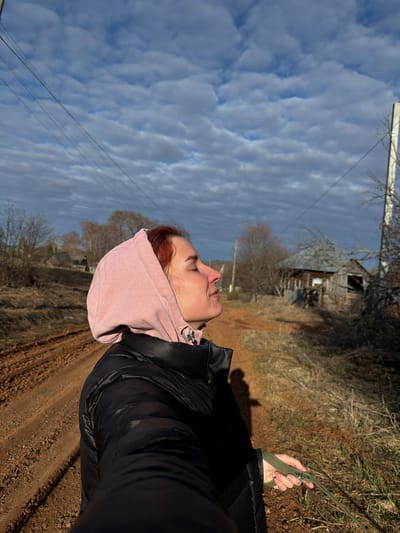 A medium shot, taken outdoors in Заболотово, Russia, shows a person from the waist up, their face tilted skyward. The person wears a black puffy jacket and a pink hooded sweatshirt. Their red hair is visible at the edge of the hood. They hold a green leash in their right hand. The background features a dirt road with tracks, dry grass, a wooden, dilapidated building, bare trees, and a sky filled with textured, layered clouds. Overhead wires cut across the upper part of the sky.