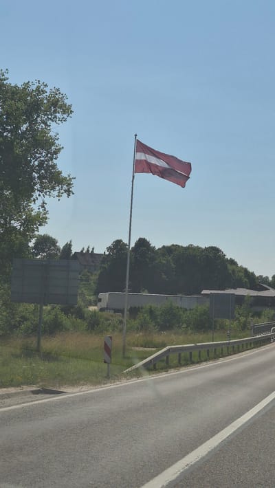 The image depicts a roadside scene under a clear blue sky. A Latvian flag flies on a flagpole to the right of center. In the background, there is a dense line of trees, and beyond that, some buildings are visible. A white truck is parked on the side of the road near the trees. A metal guardrail runs along the edge of the road in the foreground. The road itself is paved and appears to be a highway or major road. The overall environment suggests a rural or semi-rural setting. The location is the city of Smėlynė, Lithuania.