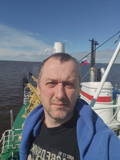 The photograph shows a man, presumably in his late 40s or early 50s, with short grey hair and stubble, wearing a black t-shirt and a blue towel draped over his shoulders. He is positioned on a boat or ship, possibly in Veregonets, Russia, as indicated by the Russian flag visible in the background. The backdrop features a body of water, possibly a river or sea, with land visible in the distance. The sky is partly cloudy. The vessel appears to be functional, given the presence of deck equipment and railings. The location may be inferred due to the flag and boat, possibly Veregonets, Russia.