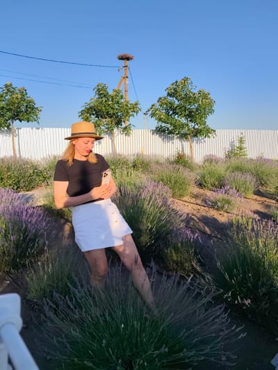 A young woman wearing a straw hat, a black t-shirt, and a white skirt stands in a field of lavender. She is holding a smartphone and appears to be taking a photo or recording a video. The woman has blonde hair and is wearing red earrings. She is leaning against a dark surface, possibly a rock or a planter.
Behind her, there is a white corrugated metal fence. Several trees with green foliage are planted in front of the fence. In the background, a wooden pole with a stork's nest is visible against a clear blue sky. The sun is low in the sky, casting long shadows, suggesting it is either early morning or late afternoon. The overall scene depicts a pleasant, outdoor setting in a rural or semi-rural area, with the lavender field being a prominent feature. The woman's posture and engagement with her phone suggest she is enjoying the moment and documenting her experience. There are no visible text elements in the image.