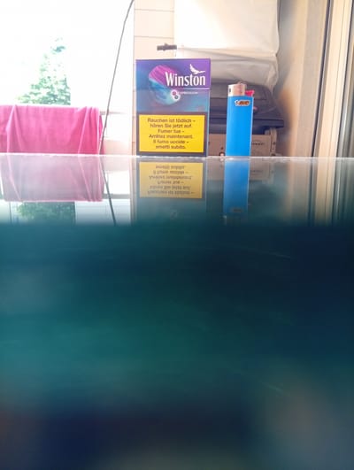 The image depicts a close-up, slightly angled shot of a glass surface reflecting items on a balcony. The subjects visible on the balcony include a pack of Winston cigarettes, a blue BIC lighter, and a pink towel draped over a railing. The Winston pack is upright and features the brand name prominently. The blue lighter stands next to it. The pink towel is partially visible on the left. The glass surface below shows a distorted reflection of these items and the surrounding environment, including greenery. The lower portion of the image is dominated by the deep teal and dark blue hues of the water or liquid in a container, suggesting this glass surface is part of a tank or aquarium. The background is bright, indicating outdoor lighting, with hints of trees and building structures. The location is identified as Langenthal, Switzerland.