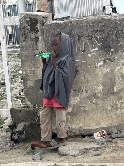 A young child, likely a girl, stands in front of a rough concrete wall in a gritty urban environment, possibly Lagos, Nigeria. She is wearing a dark grey hijab that covers her head and shoulders, a red top underneath, and loose-fitting tan trousers with a faded emblem on the right leg. Her feet are clad in light green slide sandals. The child is holding a small, brightly colored green packet, possibly food, up to her mouth, suggesting she is eating or drinking from it.
The background features the textured and weathered concrete wall, marked with faded graffiti and peeling posters. To the left, a portion of a white metal fence and a concrete structure are visible, with debris and rubble scattered around the ground. To the right of the child, on the ground, lies an empty, crumpled red and white box that appears to be for a product labeled "CREAM." The overall scene depicts a scene of everyday life in a densely populated urban area, with signs of wear and tear on the infrastructure.
