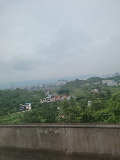 The image depicts a cityscape viewed from a slightly elevated position, likely from a vehicle, overlooking rolling green hills and buildings. In the foreground, a concrete barrier with a textured, weathered surface runs horizontally across the bottom of the frame. Beyond the barrier, the landscape unfolds with lush green foliage covering the hills.

Nestled within the greenery are various structures. Several residential buildings, characterized by tiled roofs in shades of blue and brown, are scattered across the slopes. Some appear to be multi-story houses. Further in the distance, a more developed area is visible with clusters of modern buildings, including apartment complexes and office buildings, showcasing a mix of architectural styles with balconies and windows. The tallest structures appear to be in the background, suggesting a more urbanized zone further away.

The sky above is overcast, with a uniform grey cloud cover that diffuses the light, creating a soft, even illumination across the scene. The overall impression is one of a suburban or semi-urban area with a blend of natural landscape and human development, possibly in the Yufengshan area of China, given the context. There are no discernible individuals or active interactions visible in the frame.