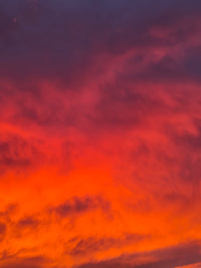 The image depicts a dramatic sunset sky over Campochiaro, Italy. The sky is filled with dense clouds painted in vibrant hues of deep purple, intense red, and fiery orange. Wisps of darker clouds are visible, creating texture and depth against the bright backdrop. The overall impression is one of a powerful and colorful atmospheric display. There are no people, subjects, or objects clearly discernible in the image, and no actions or interactions are taking place. The environment is solely the sky.