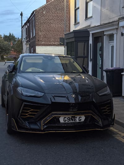 A customized matte black Lamborghini Urus, bearing the United Kingdom registration plate "G9NE G," is parked on an asphalt residential street in Boston, United Kingdom. The vehicle features prominent gold or yellow accent lines along its front bumper, grilles, and lower lip. The car's front is oriented towards the viewer, slightly angled to the left, showcasing its aggressive styling, visible Lamborghini emblem, and intricate headlight design.

The street itself consists of dark asphalt, with a visible curb and paved sidewalk to the right. In the background, a row of terraced houses lines the street. The houses on the left are primarily red brick, with one exhibiting a painted white lower section. A utility pole with overhead power lines is visible against a cloudy or twilight sky above these houses. Towards the right, the houses are painted in lighter tones, possibly light grey or white, with dark-framed windows. One dwelling features a modern black bay window extension, while another displays a decorative white lintel above its entrance. A black domestic refuse bin with a purple lid is positioned on the sidewalk to the far right. No individuals are visible in the scene.