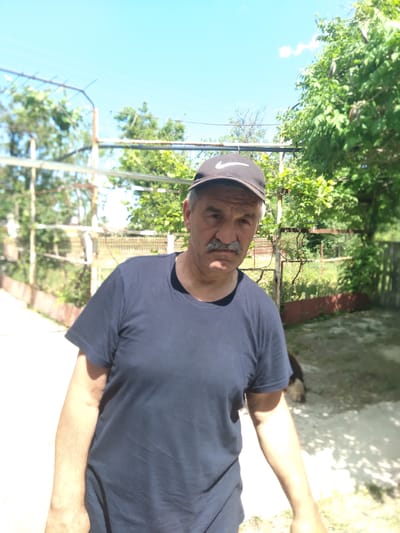 This media file captures a portrait of a middle-aged to older man outdoors on a clear, sunny day. The man is looking directly at the camera with a neutral expression. He has a prominent gray mustache and is wearing a dark blue short-sleeved t-shirt and a dark baseball cap. A white Nike swoosh logo is clearly visible on the front of his cap. His right arm is extended to the side of the frame.
The background consists of lush green foliage from trees or vines, suggesting a garden or a natural, overgrown area. A rusty-looking chain-link fence or wire trellis structure is visible behind him, further indicating an outdoor, possibly rural or suburban, setting. The sky above is bright blue with a few small white clouds. The ground appears to be a mix of dirt and concrete, with some shadows cast by the surrounding vegetation. A dark-colored dog is partially visible in the upper right background, near a light-colored wall. The scene is illuminated by bright daylight, suggesting it is midday. The location context is Журавское сельское поселение, Ukraine.