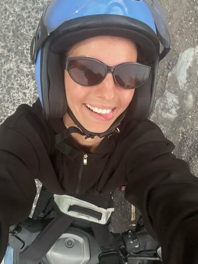 A person is riding a motorcycle in Loloan, Indonesia, and sticking their tongue out at the camera. The person is wearing a blue helmet, sunglasses, and a black hoodie. The motorcycle is black and silver. The background is a paved road with gravel and dirt.