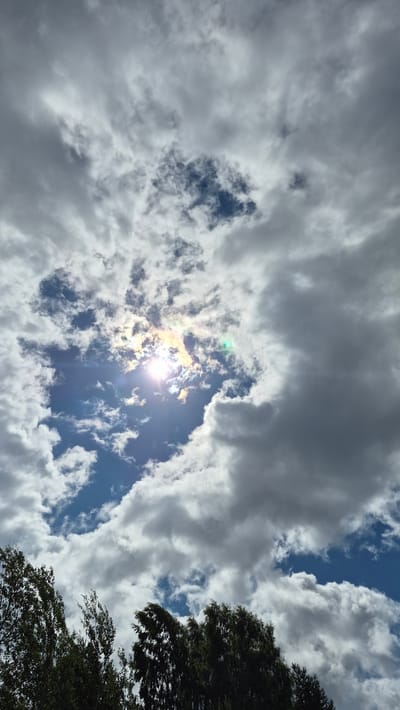 This is a vertical, medium-angle shot of a partly cloudy sky, with the sun shining brightly through a break in the clouds. The sun is near the center of the frame, radiating light in a way that creates a lens flare effect with rainbow hues. The clouds are a mix of bright white and darker gray, with some appearing wispy and others more dense. Below the sky, the tops of dark green trees are visible, their leaves appearing slightly blurred as if from wind or movement. The overall impression is of a dynamic sky on a possibly windy day. The location is Ļūbaste 0001, Latvia. There are no people or animals visible in the image.