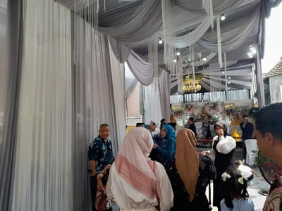 The image depicts a lively social gathering, likely a wedding reception or similar celebratory event, held within a decorated tent in Kecamatan Gempol, Indonesia. The venue is adorned with elegant white and grey fabric drapes cascading from the ceiling and forming walls, creating a festive and semi-enclosed atmosphere. White decorative fringes or string lights hang from the draped ceiling, and ornate chandeliers with multiple bulbs illuminate the space.
At the far end of the tent, a focal point resembling a stage or elaborate backdrop is visible. It is decorated with abundant floral arrangements in shades of white, pink, and purple, interspersed with various ornaments, suggesting a space for a bridal couple or honored guests.
Numerous guests are present, standing and mingling throughout the tent. The attendees primarily appear to be adults, with a few children visible. Many women are wearing hijabs in various colors, including blue, brown, and light pink. Men are dressed in patterned shirts, with one prominent figure on the left wearing a dark blue patterned shirt, possibly batik. People appear engaged in conversation and observing the event. A young child with white bows in their hair is partially visible in the foreground on the right. A large fan is also visible on the right, suggesting the need for cooling in the outdoor or semi-outdoor setting.
The bright illumination within the tent indicates the event is taking place during the daytime. The general attire and decorative elements are consistent with cultural celebrations in Indonesia. There is no legible text visible in the image.