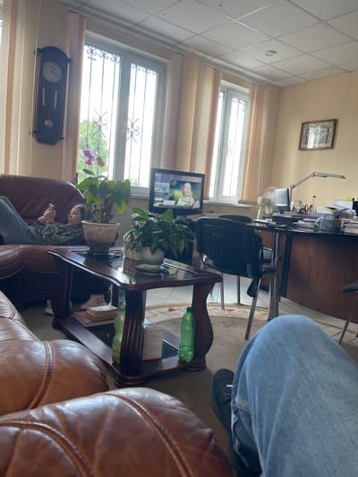 In a room in Makariv, Ukraine, a young girl with dark hair lies on a brown leather couch, looking at her phone. She wears a sparkly black top and blue jeans. In front of the couch, a dark wood coffee table holds potted plants, a book, and two plastic bottles. A large grandfather clock hangs on the wall near a window with vertical blinds. A desk with a computer, fan, and paperwork is visible in the background. Two brown leather couches are visible in the foreground.