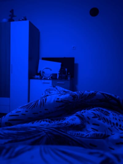 The scene depicts a room in Nadarevo, Bulgaria, illuminated predominantly by a monochromatic blue light. The foreground is occupied by a bed covered with rumpled bedsheets and a duvet, featuring a dark, organic leaf-like pattern against a lighter background. No individuals are visible. In the midground, to the left, stands a light-colored, tall wardrobe or cabinet with a vertical handle. To its right, a lower dresser or chest of drawers holds various objects on its surface, including a round mirror with a stand, a rectangular item (possibly a screen or board) leaning against the wall, and other indistinct items. A dark, rectangular object, potentially a television, is visible behind the dresser, mounted on the wall or standing. The wall itself is a plain, light color, appearing deep blue under the specific lighting. A single electrical outlet is visible on the mid-right section of the wall. High on the wall, towards the center, a small, dark, circular object is affixed.